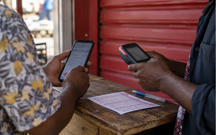 close adult Tanzanian hands comparing phone confirmation with agent terminal beside paper betting coupon, red shutter backdrop; using Airtel Money for an account payment. Illustrative generated scene.