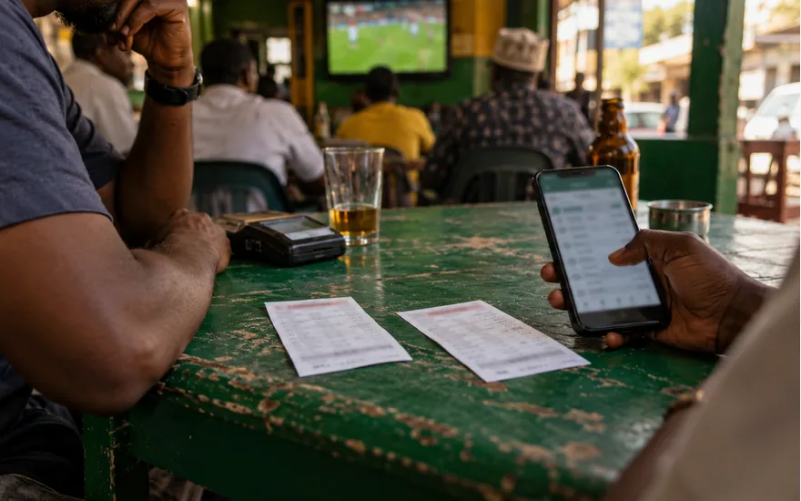Two anonymous printed betting coupons and a phone with blurred ticket history on a worn green sports cafe table in Tanzania; how to use bet id in betpawa. Illustrative generated photograph, not an operator screenshot.