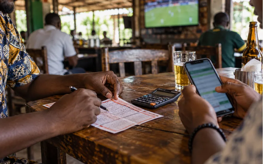 adult hands comparing printed betting coupon with smartphone and small calculator at a Tanzanian sports bar table; checking a published bonus. Illustrative generated scene.