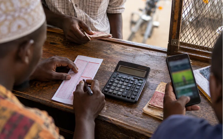 overhead on a worn wooden betting kiosk counter: calculator, paper bet slip, anonymous cash and blurred casino phone; checking a published bonus. Illustrative generated scene.