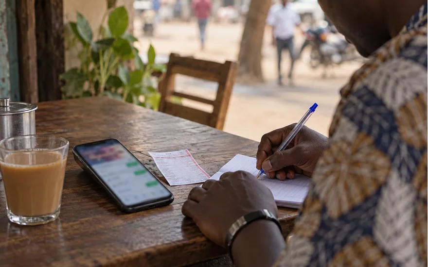 adult Tanzanian bettor writing transaction reference on paper next to smartphone with blurred support chat and betting ticket; getting an account or payment problem investigated. Illustrative generated scene.