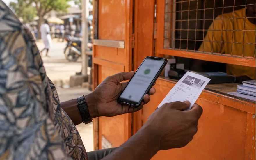 orange-painted unbranded Tanzanian mobile money kiosk, adult hands holding phone with blurred deposit confirmation and sports ticket; using Halo-Pesa safely in the cashier. Illustrative generated scene.