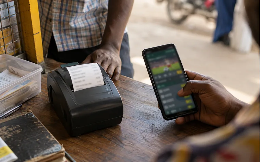 overhead of small agent terminal beside adult dark-skinned hand and blurred betting phone, neutral Tanzanian kiosk counter; using Halo-Pesa safely in the cashier. Illustrative generated scene.