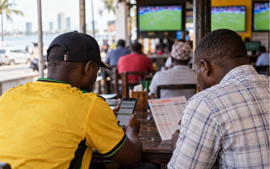 Two adult Tanzanian football fans from behind looking at a phone and printed fixture sheet in a daylight Dar es Salaam sports cafe; betpawa jackpot. Illustrative generated photograph, not an operator screenshot.