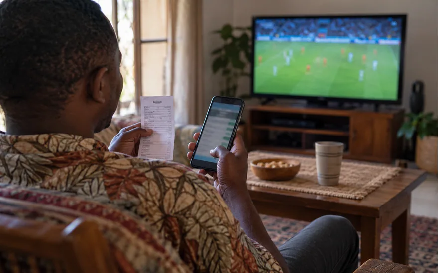 over shoulder adult Tanzanian man viewing smartphone betting slip during football broadcast, sober candid atmosphere; understanding live-bet acceptance. Illustrative generated scene.