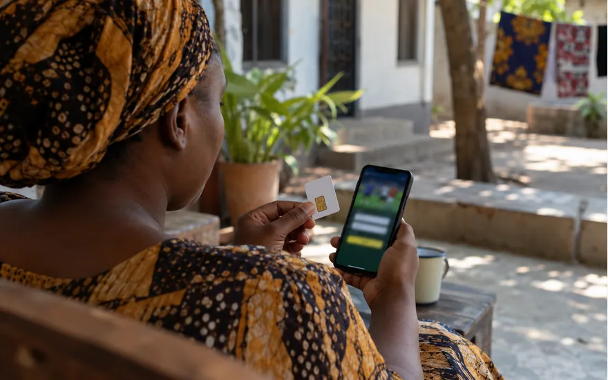 adult woman from behind holding SIM card packaging next to a phone showing blurred betting login, shaded Dar es Salaam courtyard; signing in to an existing account. Illustrative generated scene.