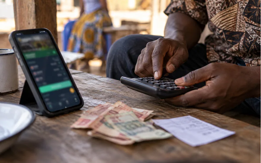adult Tanzanian hands using small calculator beside phone with blurred betting deposit amount screen and unidentifiable cash; checking the smallest permitted cashier payment. Illustrative generated scene.