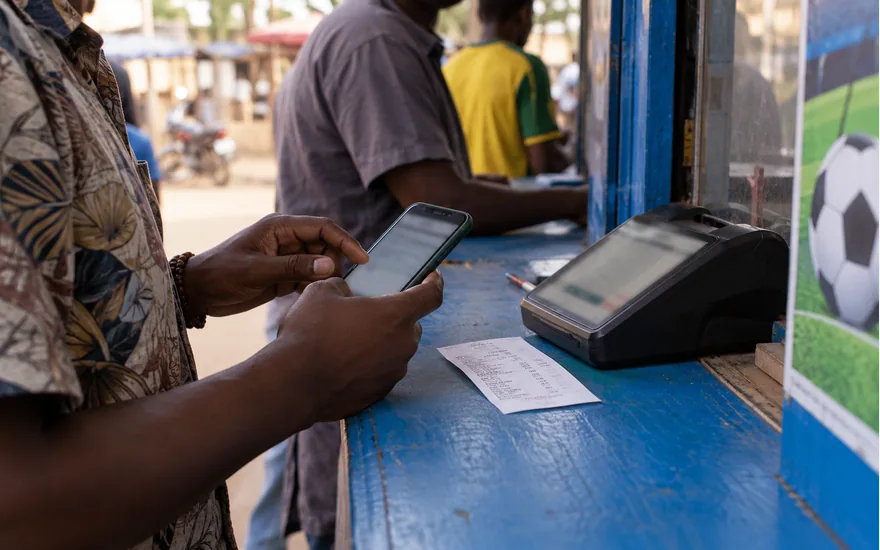 Tanzanian adult hands on mobile money phone beside terminal and unreadable sports bet ticket at blue-painted kiosk counter; using the Mixx by Yas payment route. Illustrative generated scene.