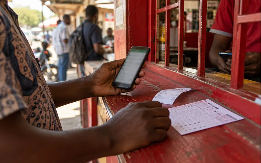 adult Tanzanian hand holding phone at red accented mobile money counter beside unreadable transfer receipt, betting coupon; using the Vodacom M-Pesa payment route. Illustrative generated scene.