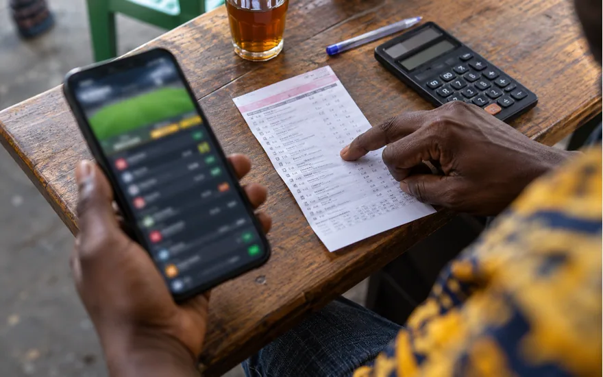 close overhead phone with blurred casino coupon screen and adult dark-skinned hand pointing to unreadable ticket beside calculator; verifying a no-deposit claim. Illustrative generated scene.
