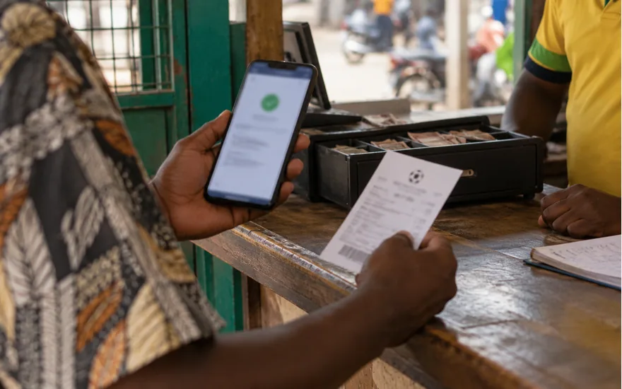 adult Tanzanian hand presenting phone with blurred mobile payment confirmation at local agent counter, cash drawer in background; moving money between your wallet and account. Illustrative generated scene.