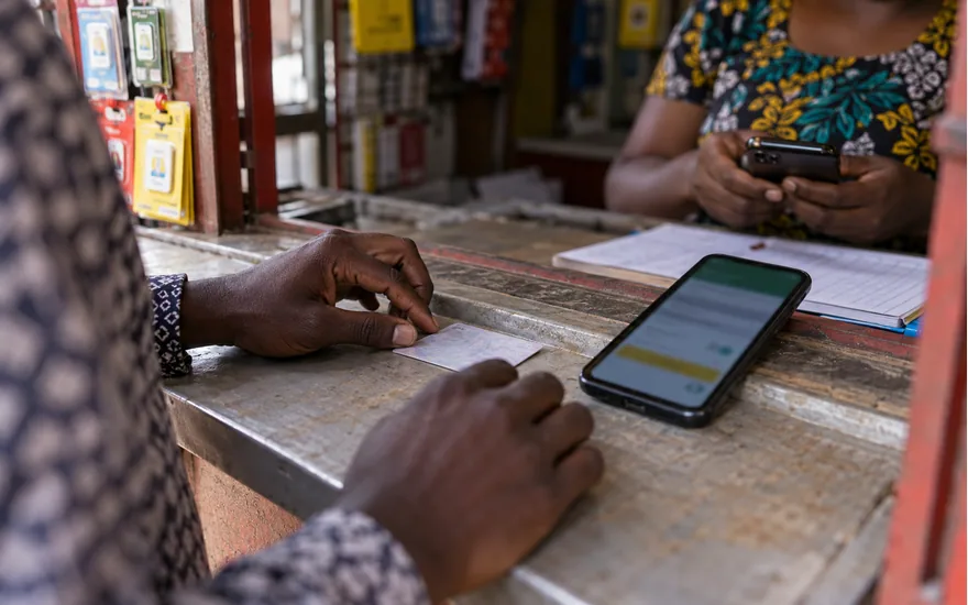 Tanzanian adult hands placing an identity card face down beside smartphone with blurred betting signup form, at a SIM counter; opening a Tanzania account. Illustrative generated scene.