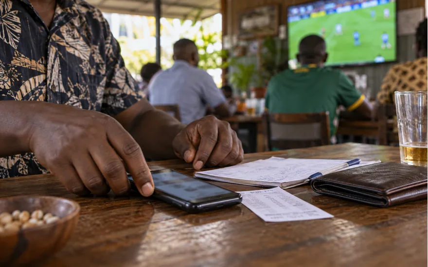 adult Tanzanian hands placing phone with blurred betting slip face down beside closed wallet and a paper budget at sports cafe; protecting money and taking a break. Illustrative generated scene.