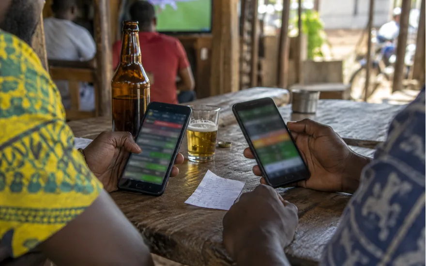 two smartphones side by side with different blurred betting screens and handwritten anonymous betting slip on Tanzanian bar table; deciding what this review actually proves. Illustrative generated scene.