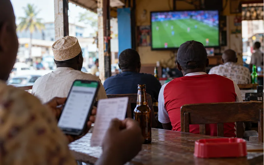football television in small Dar es Salaam sports bar with adult spectators from behind, foreground phone with blurred sports coupon; reading and placing a sports coupon. Illustrative generated scene.