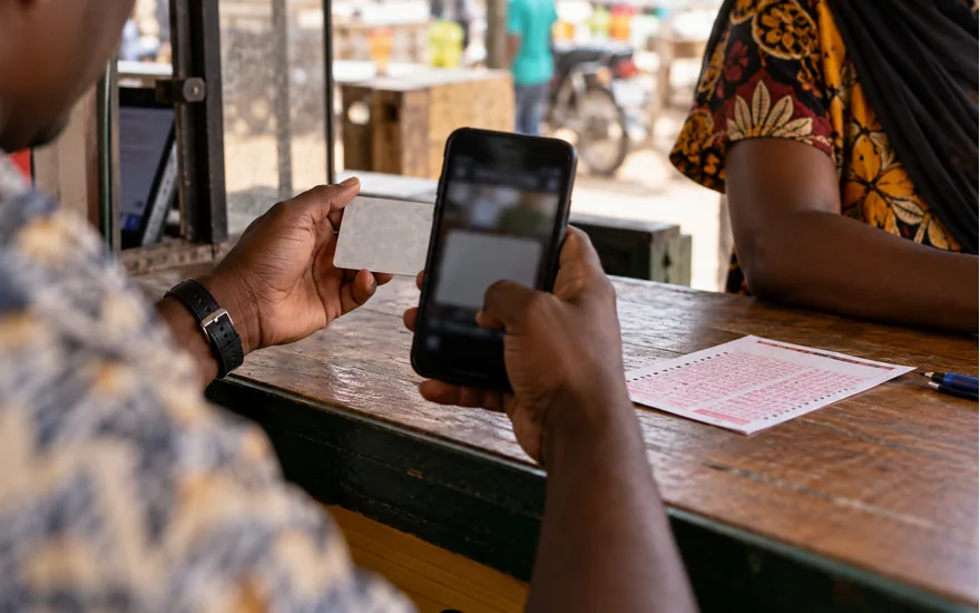 adult Tanzanian hands holding phone camera over blank back of identity card, betting coupon on counter, no visible identity data; completing an identity check. Illustrative generated scene.
