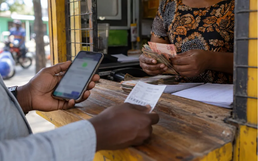 Tanzanian mobile money kiosk agent hands counting cash behind counter beside customer phone with blurred betting withdrawal confirmation; withdrawing to your own mobile wallet. Illustrative generated scene.